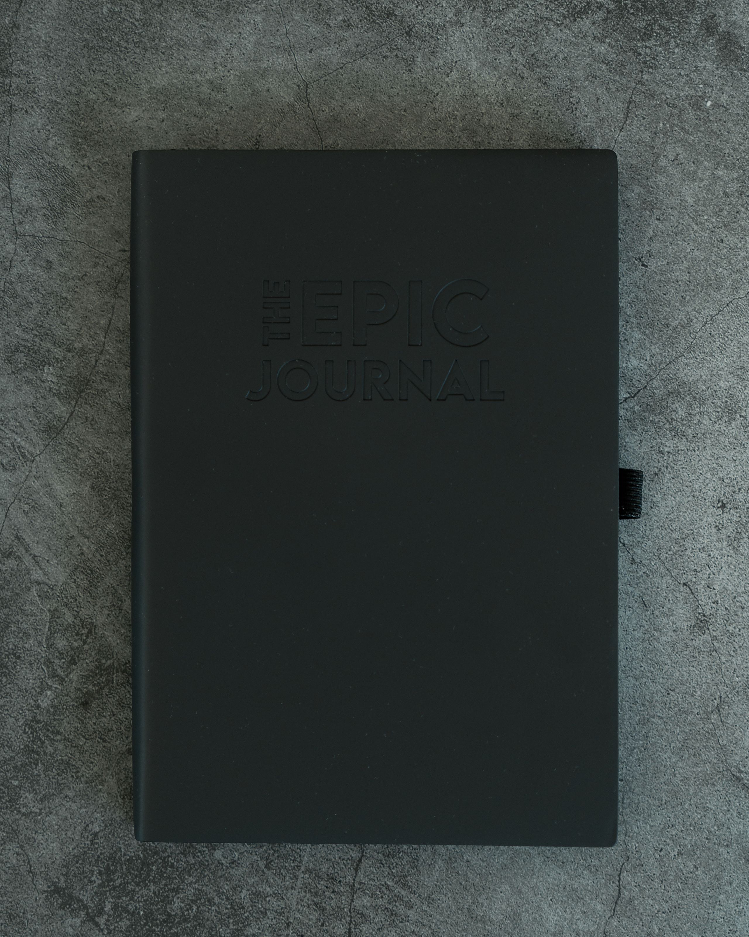 The Epic Journal in matte black vegan leather, closed and embossed, resting on a concrete surface